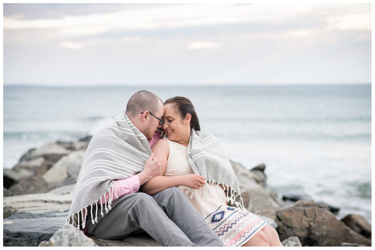 Couple sitting on rocks at the beach while wrapped in blanket at sunset