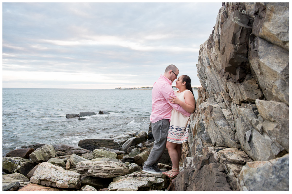 Engaged couple looking at each other on rocks next to ocean in New England